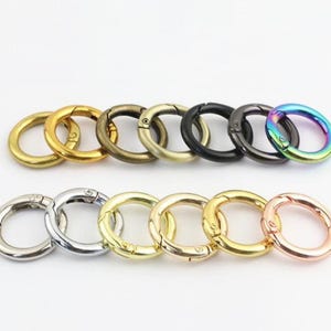 May include: A collection of thirteen circular spring-loaded metal gate rings arranged in two rows. The rings feature various metallic finishes including gold, silver, bronze, matte black, gunmetal, rose gold, and a multi-coloured iridescent finish.