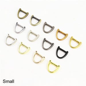 May include: A collection of small D-rings in various metallic finishes including gold, silver, bronze, gunmetal, and black. Each D-ring features a curved bottom and a straight top bar, arranged in a scattered pattern on a plain white background.
