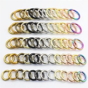May include: A collection of circular metal spring gate rings arranged in five horizontal rows. The rings feature various metallic finishes including gold, bronze, black, silver, rose gold, and a multi-coloured iridescent rainbow gradient.