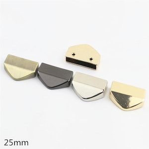 May include: Five metal bag edge connectors measuring 25mm, featuring a pointed, shield-like shape. The set includes finishes in antique brass, matte black, polished silver, bright gold, and a textured gold piece with two small screw holes.