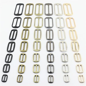 Può includere: Una collezione di cursori rettangolari in metallo per cinghie disposti in una griglia. I cursori presentano una barra centrale e sono disponibili in varie finiture metalliche, tra cui nero opaco, ottone antico, oro spazzolato, canna di fucile, argento e oro.