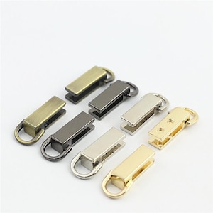 Puede incluir: Un conjunto de ocho tiradores de cremallera rectangulares de metal dispuestos en dos filas. El set incluye varios acabados metálicos como latón antiguo, bronce, plata y tonos dorados, cada uno con un pequeño anillo circular adjunto.