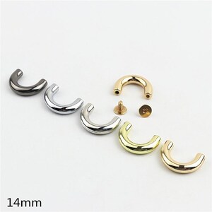 Op de afbeelding: Een verzameling van 14 mm C-vormige metalen onderdelen in diverse metallic afwerkingen, waaronder gunmetal, zilver en goud. De set bevat gebogen stukken met gladde, gepolijste oppervlakken en kleine schroefbevestigingen met schroefdraad.