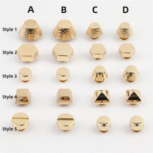Può includere: Una collezione di borchie e bottoni in metallo dorato disposti in una griglia etichettata A, B, C, D e Style da 1 a 5. Gli articoli includono forme coniche testurizzate, pezzi esagonali lisci, dischi circolari piatti, piramidi quadrate e teste di vite a taglio.