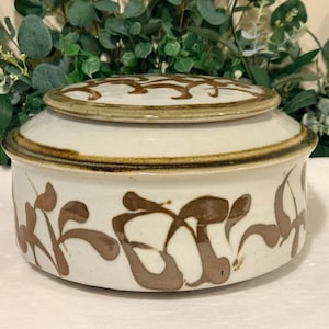 May include: A round ceramic lidded container with a cream-colored base and a brown hand-painted abstract leaf pattern. The lid features a matching brown abstract design, and the rim of the lid and the upper edge of the container are accented with a dark brown band.