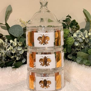 May include: A three-tiered clear glass canister set with a matching lid. Each of the three cylindrical glass sections features a white rectangular label with a gold metallic fleur-de-lis emblem and vertical gold stripes on the sides.