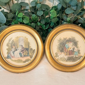 May include: Two circular wall plaques featuring ornate gold frames and central illustrations. The left plaque depicts a group of figures in a garden setting, while the right plaque shows a scene with multiple figures in a wooded landscape.