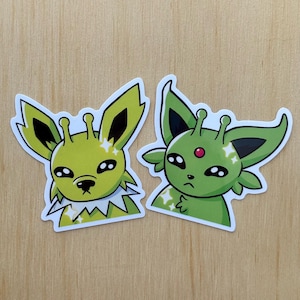May include: Two die-cut stickers featuring stylised green creatures with large ears, antennae, and dark eyes. The left sticker shows a creature with a white collar, while the right sticker depicts a creature with a small red gem on its forehead.