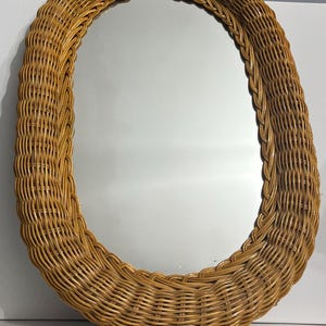 May include: An oval-shaped wall mirror featuring a thick, light brown woven wicker frame. The mirror has a smooth, reflective glass surface surrounded by a detailed, braided rattan border that creates a textured, natural appearance.