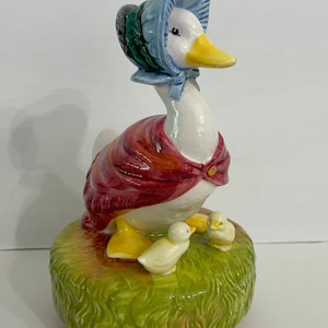 May include: A ceramic figurine of a white goose wearing a blue bonnet and a red shawl, standing on a green textured base. Two small yellow ducklings are positioned at the feet of the goose on the grass-like surface of the round base.