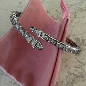 以下が含まれることがあります： 透明で輝く石がちりばめられた、ヘビのようなセグメントデザインのシルバー調のオープンブレスレット。ブレスレットは、お揃いのピンクの紐が付いた柔らかいピンクのベルベット製巾着袋の上に置かれています。