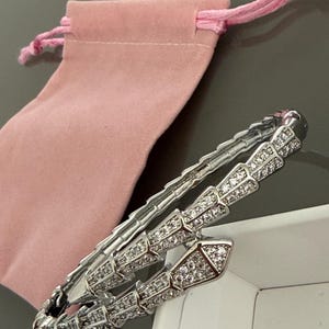 May include: A silver-toned wrap bracelet featuring a segmented, snake-like design encrusted with numerous small, clear, sparkling stones. The bracelet is accompanied by a soft, light pink fabric drawstring pouch with matching pink ties.