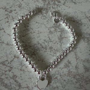 May include: A silver-toned beaded bracelet featuring a polished heart-shaped padlock charm. The bracelet includes a lobster claw clasp and a small circular tag stamped with 925. The heart charm also features the engraved text 925 on its surface.