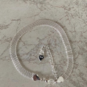 Puede incluir: Una pulsera de cadena de malla plateada con una textura tejida. La pulsera incluye una cadena extensora ajustable con un pequeño dije en forma de corazón que cuelga del extremo. El dije tiene grabado claramente el texto 925.