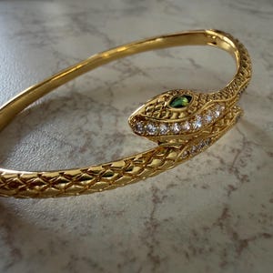 Könnte beinhalten: Ein goldfarbener Scharnierarmreif in Schlangenform. Die Metalloberfläche weist ein strukturiertes Schuppenmuster auf. Der Schlangenkopf ist mit klaren weißen Kristallen am Kiefer und einem grünen Stein als Auge verziert.