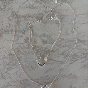 May include: A silver-toned jewelry set consisting of a necklace and a bracelet, both featuring a delicate box chain and a polished open heart pendant. Each piece includes a lobster claw clasp and a small extender chain with a 925 stamp visible.