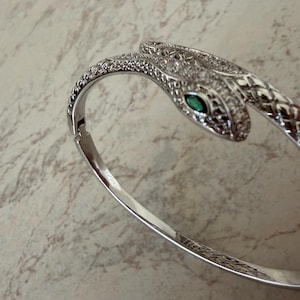 May include: A silver-toned hinged bangle bracelet shaped like a snake. The snake head features a marquise-cut green stone for an eye and is encrusted with small clear stones along the top surface, which continues down the textured body of the snake.