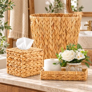 May include: A set of woven water hyacinth bathroom accessories including a tall cylindrical waste basket, a square tissue box cover with white tissues, and a rectangular tray holding a roll of toilet paper and a small white floral arrangement.
