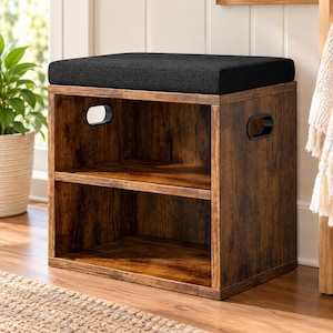 May include: A rectangular storage ottoman featuring a dark brown wood grain finish, a black upholstered seat cushion, and two open shelves. The side panels include integrated oval-shaped cutouts that serve as handles for easy movement.