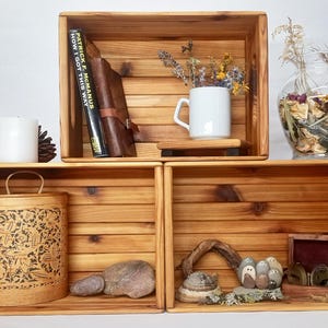 May include: Three rectangular wooden crates stacked to form shelving. The top crate holds a book titled How I Got This Way, a white mug, and dried flowers. The bottom left crate contains a patterned birch bark container. The bottom right crate holds stones and a small wooden box.