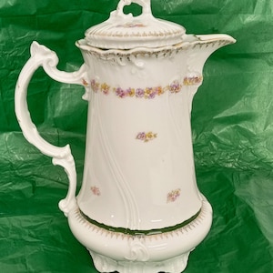 May include: A white porcelain coffee pot featuring a delicate floral pattern of small purple and yellow flowers. The pot has an ornate handle, a matching lid with a decorative finial, and a dark green band near the base with gold trim accents.