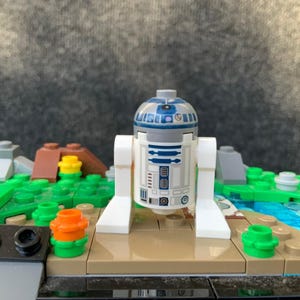 May include: A white and blue R2-D2 droid minifigure with a domed head and printed mechanical details on its cylindrical body, standing on a tan base plate surrounded by green, brown, and blue plastic building blocks on a textured surface.