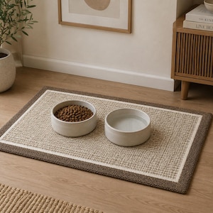 May include: A rectangular pet feeding mat with a textured beige centre and a dark brown border. Two round, light-coloured ceramic bowls sit on the mat, one filled with brown dry kibble and the other filled with clear water for a pet.