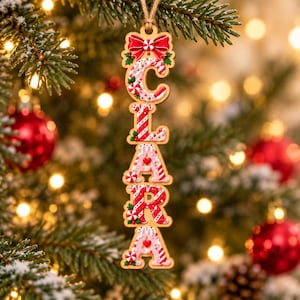 May include: A vertical wooden Christmas ornament featuring the name CLARA spelled out in candy cane-themed letters. Each letter is decorated with red and white stripes, peppermint swirls, and small red hearts, topped with a red bow.
