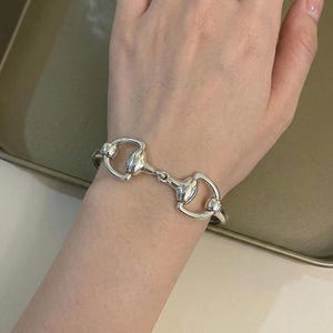 May include: A polished silver-tone bracelet featuring a central horsebit-inspired metal link design. The bracelet consists of a smooth, curved metallic bar that connects two symmetrical, rounded loops, creating a continuous, sleek, and reflective accessory.