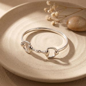 Könnte beinhalten: Ein polierter Silberarmreif mit einem zentralen Trensen-Design. Das kreisförmige Band ist glatt und reflektierend und zeigt eine klassische, vom Reitsport inspirierte Metallverbindung, die die beiden Seiten des starren Reifens verbindet.