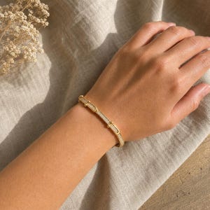 Könnte beinhalten: Ein goldfarbenes Armband bestehend aus einer Reihe rechteckiger Glieder, die mit kleinen, klaren, funkelnden Steinen besetzt sind. Das Armband hat eine polierte Oberfläche und ein strukturiertes Design mit abgerundeten Metallakzenten an den Verbindungsstellen.