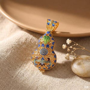 Puede incluir: Un colgante de metal dorado con forma de calabaza, que presenta un intrincado trabajo de filigrana calada y detalles de esmalte de colores en azul, verde y blanco. El diseño incluye un símbolo circular central y una reasa decorativa.