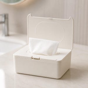 Può includere: Una scatola dispenser rettangolare in plastica color bianco sporco con coperchio incernierato aperto. Una singola salvietta bianca è parzialmente estratta attraverso l'apertura circolare centrale sulla parte superiore del contenitore opaco.