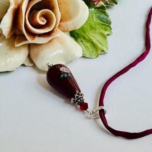 Puede incluir: Un colgante de cristal en forma de lágrima de color granate oscuro con un pequeño diseño floral pintado a mano. El colgante está decorado con una tapa de metal plateado, una pequeña cuenta roja facetada y una anilla plateada en un cordón.