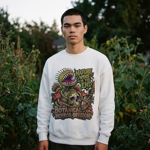 May include: A cream-colored crewneck sweatshirt featuring a graphic print of a Venus flytrap, a skull, mushrooms, and a garden trowel. The text reads, "NATURE WINS IN THE END" and "BOTANICAL HORROR DIVISION" in a dark green and brown color palette.