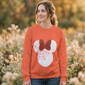 Puede incluir: Una sudadera naranja de manga larga con una gran silueta blanca en forma de cabeza de ratón. El diseño incluye un patrón de telaraña en una oreja y un lazo de color naranja oscuro con purpurina centrado entre las orejas.