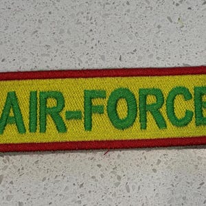 May include: Embroidered patch with the words "AIR-FORCE" in green block letters on a yellow background. The patch has a red border.