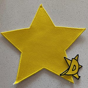 May include: A large, yellow star-shaped patch with a smaller, stylized yellow and black design. The star has a textured surface and a yellow border. The smaller design features the letter "D".