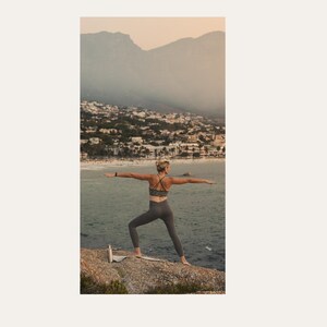 May include: A person in a dark gray sports bra with a strappy back and matching high-waisted leggings performing a yoga pose on a rocky cliff overlooking the ocean, with a small silver water bottle placed on a mat nearby against a mountain backdrop.