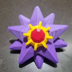 May include: A three-dimensional star-shaped object with ten purple points. Centered on the front is a yellow gear-like component with a red octagonal center piece. The object has a textured surface consistent with 3D printed plastic construction.