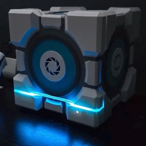 May include: A white and gray cubic device featuring a circular blue center panel with a central aperture logo. The cube has recessed geometric detailing and a bright blue LED light strip running horizontally across the front, emitting a glow.