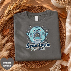 May include: A graphite heather t-shirt featuring a cartoon illustration of a light blue CT scanner with a smiling face. Below the scanner, the text reads "Scan-tastic CT Tech" in dark blue lettering, surrounded by small white star accents.