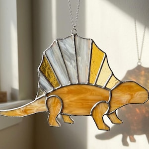 May include: A stained glass suncatcher shaped like a Dimetrodon dinosaur, featuring a body of marbled amber glass and a large dorsal sail constructed from alternating panels of translucent white and golden yellow glass, all held together by silver metal solder.