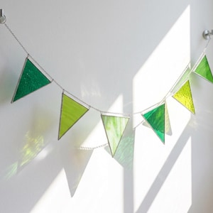 May include: A hanging garland featuring six triangular stained glass pieces in varying shades of green, ranging from light lime to deep emerald. Each glass triangle is framed in metal and connected by a silver-toned chain suspended from two wall hooks.