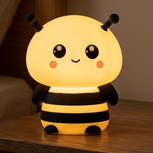 May include: A bee-shaped night light with a glowing yellow head, black and yellow striped body, two antennae, and a cartoon face featuring large black eyes, a small smile, and pink cheeks. The lamp emits a soft, warm light from its rounded head.