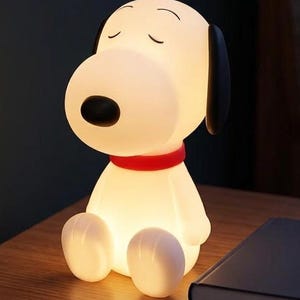 Könnte beinhalten: Eine weiße Snoopy-Charakterlampe mit glatter, matter Oberfläche, schwarzer Nase, geschlossenen Augen und einem leuchtend roten Halsband. Die Lampe strahlt ein sanftes, warmes Licht von innen aus und beleuchtet die sitzende Figur.