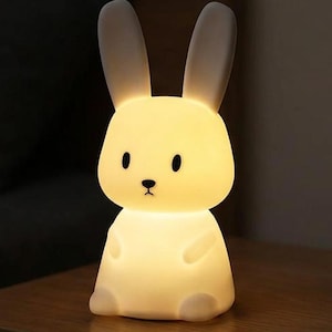 Puede incluir: Una lámpara LED blanca con forma de conejo que emite un suave resplandor amarillo cálido. La lámpara presenta orejas largas y erguidas, una cabeza redondeada y un cuerpo pequeño con ojos y nariz oscuros, creando una silueta suave e iluminada.