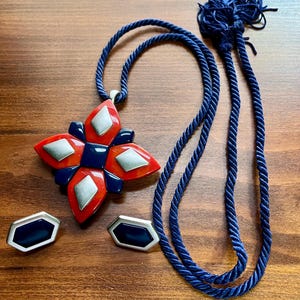 May include: A jewelry set consisting of a pendant on a dark blue twisted cord and a pair of matching earrings. The pendant features a four-pointed star shape with red, dark blue, and silver-toned diamond-shaped accents. The earrings are hexagonal.