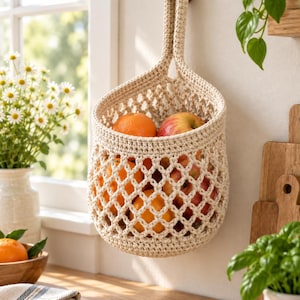 Può includere: Un cesto sospeso lavorato all'uncinetto color crema con un design a rete a rombi. Il cesto presenta un unico manico robusto appeso a un gancio da parete in legno ed è riempito con arance e mele rosse fresche per riporre la frutta.