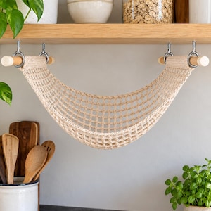 Puede incluir: Una hamaca para frutas de macramé beige con un patrón tejido en forma de rejilla, suspendida de dos varillas de madera clara. Las varillas están fijadas a la parte inferior de un estante mediante cuatro ganchos y anillos metálicos plateados.
